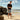 Man wearing charcoal Archies flop flops standing on a beach with large rocks and ocean in the background