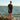 Man standing on rocks by the ocean wearing tan Archies flip flops, a black shirt and shorts.