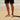Person wearing black shorts and white Archies flip-flops standing on a sandy beach with ocean waves in the background.